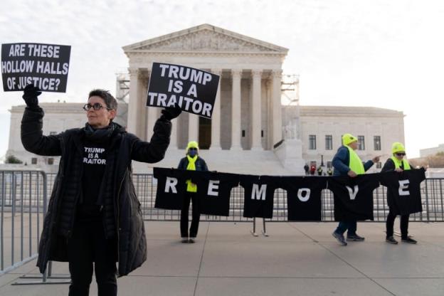 A small handful of people stand outside the steps to an august building holding signs at a demonstration. A woman holds signs that say 'Trump is a traitor' and 'Are these hollow halls of justice.'